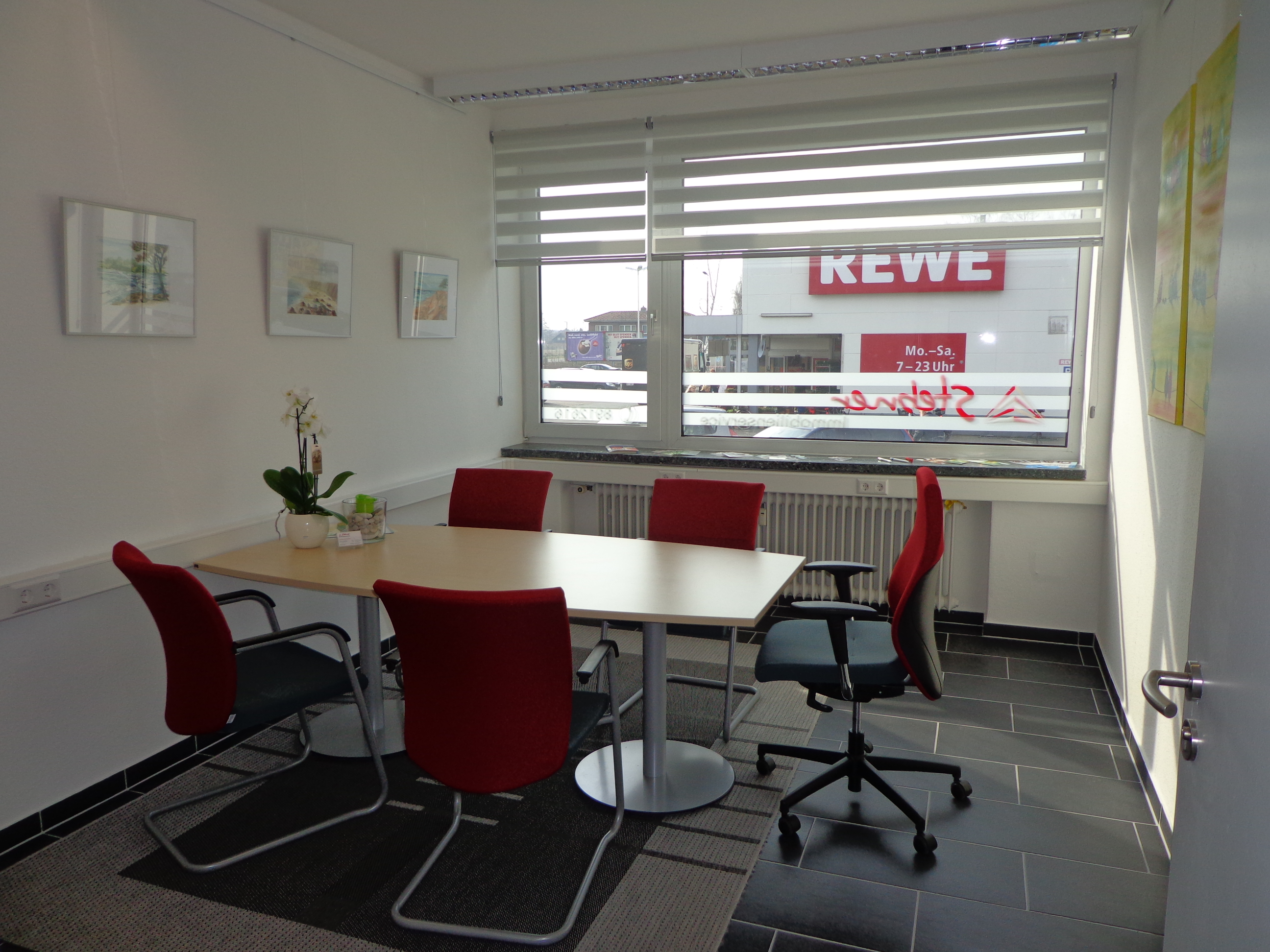Das schöne Büro von Stebner Immobilien von innen, 5 rote Stühle, ein Eckiger Tisch in der Mitte mit einer Topfpflanze darauf. Rewe ist durch das Fenster im Hintergrund zu sehen.